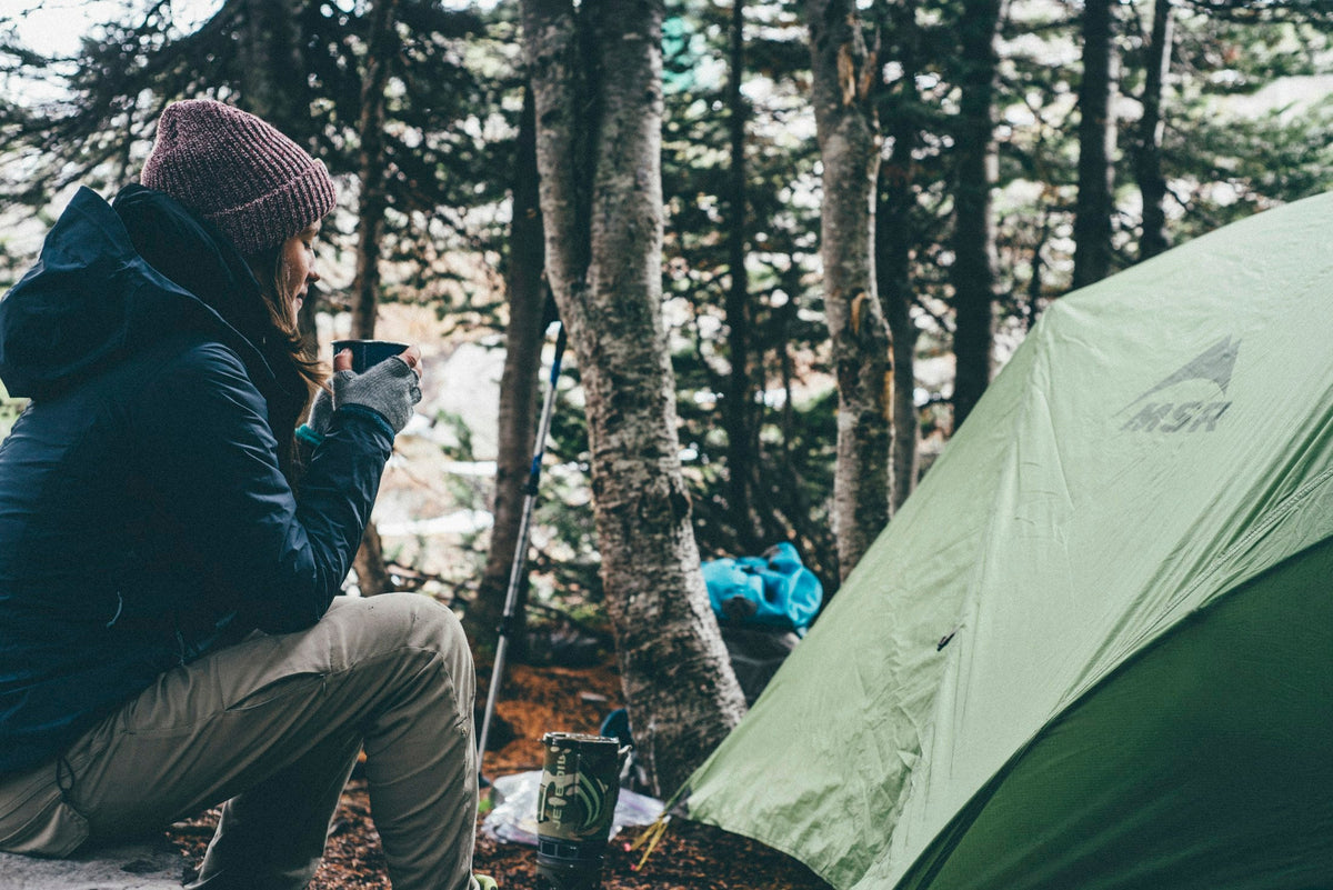 Camper holding a hot drink beside a green tent in the forest